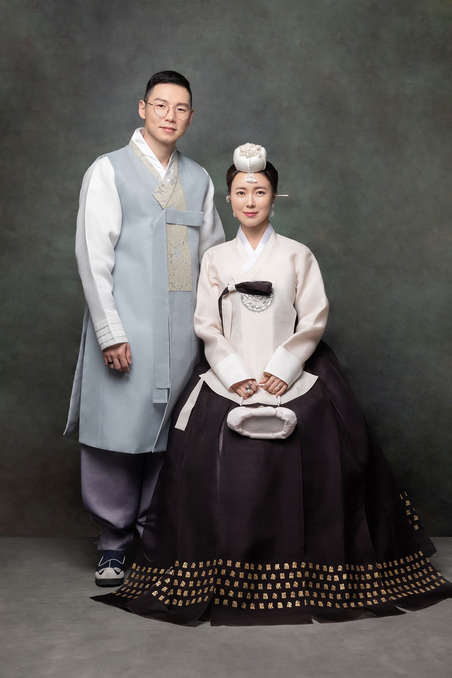 Couple wearing traditional Korean hanbok attire, man in gray jeogori and woman in white and dark brown ceremonial dress.