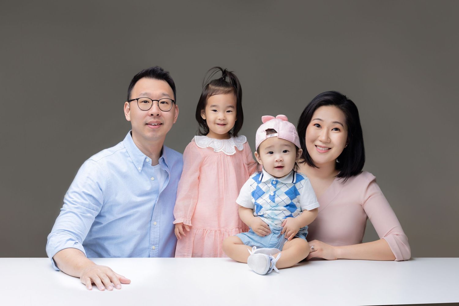 Asian family portrait with father, toddler girl, baby boy, and mother posing together at a white table.