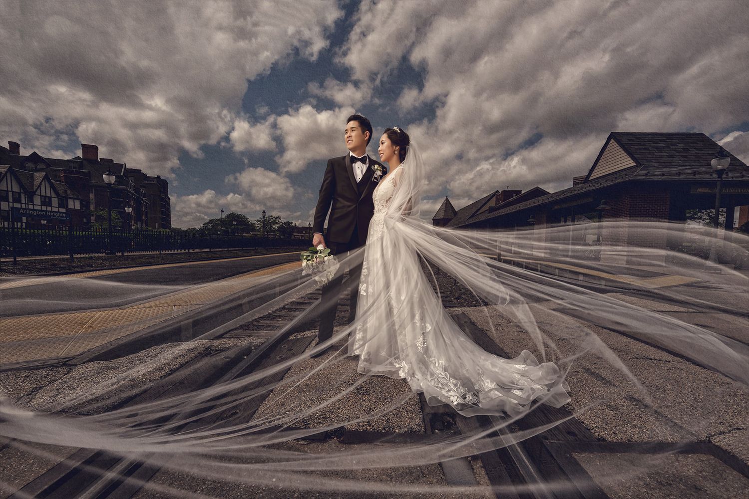Dramatic wedding photo with flowing white veil billowing in the wind against moody cloudy sky and architectural backdrop.