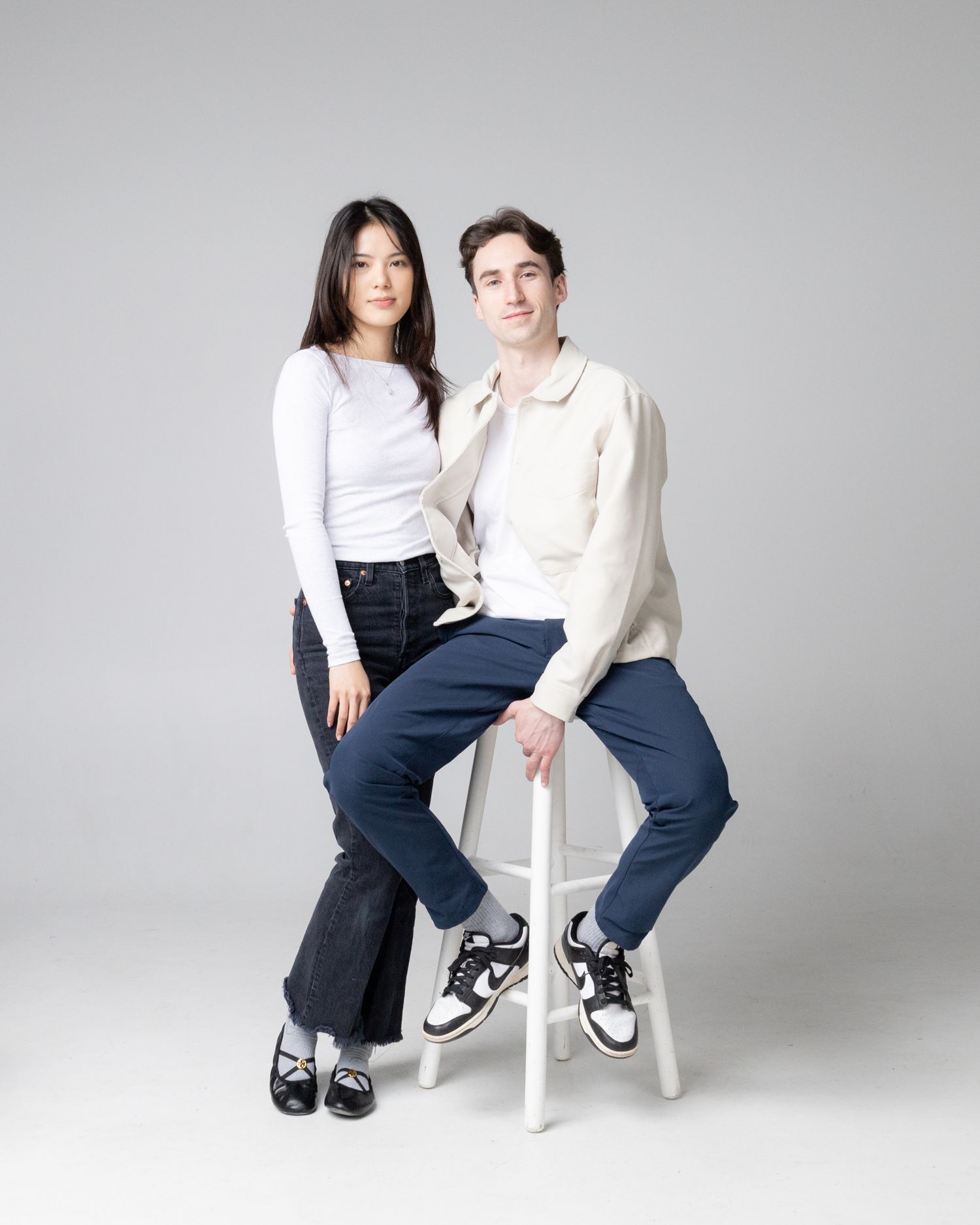 Two people in casual attire pose together on a white chair against a light gray background in various positions.