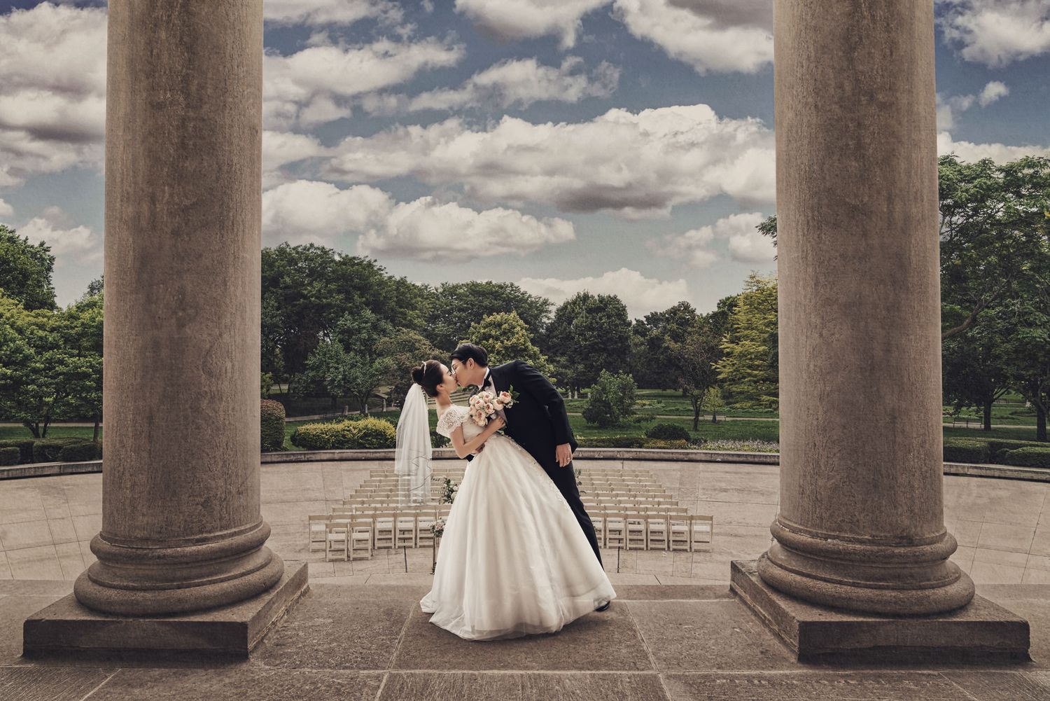 A romantic wedding moment captured between pillars at a park terrace with dramatic clouds in the sky.