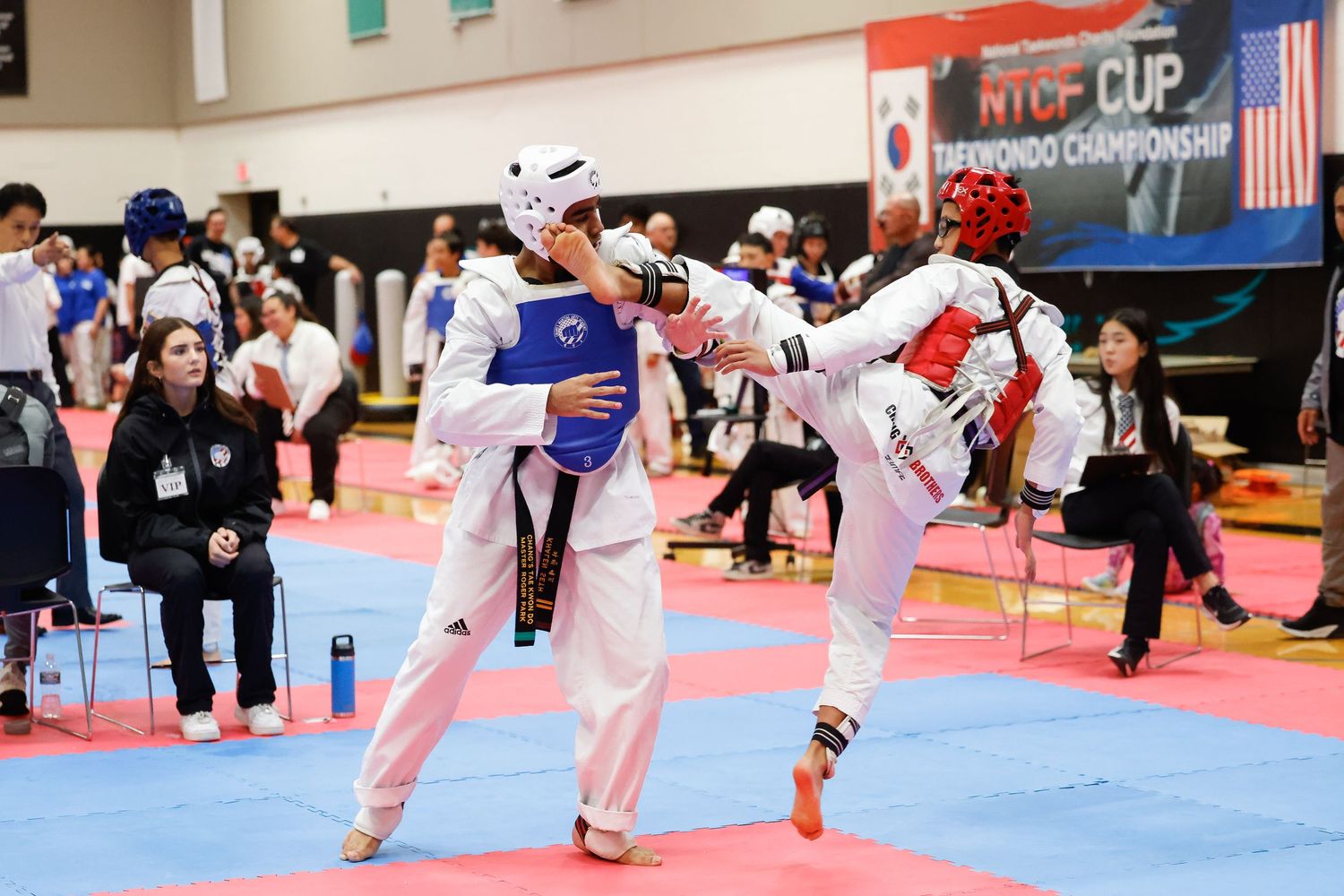 Two taekwondo competitors spar at the NTCF Cup Taekwondo Championship, one executing a high kick during the match.