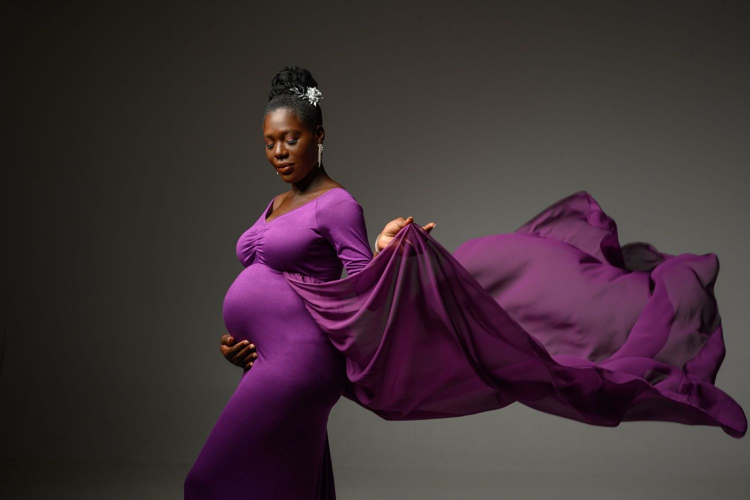 A pregnant woman in a flowing purple dress poses elegantly against a dark background with fabric billowing behind her.