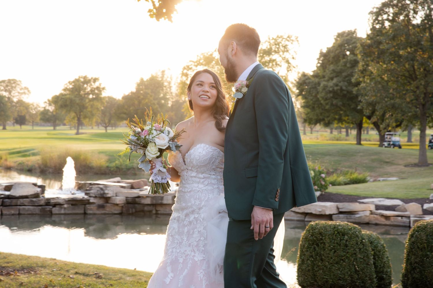 Couple in elegant wedding attire share a romantic moment at sunset by a pond with stone features and manicured gardens.