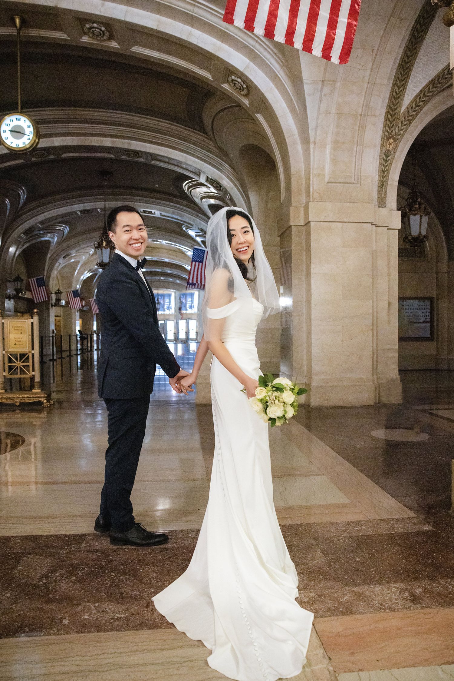Bride and groom holding hands in elegant grand hall with arched ceilings and American flags, bride in white gown with bouquet.