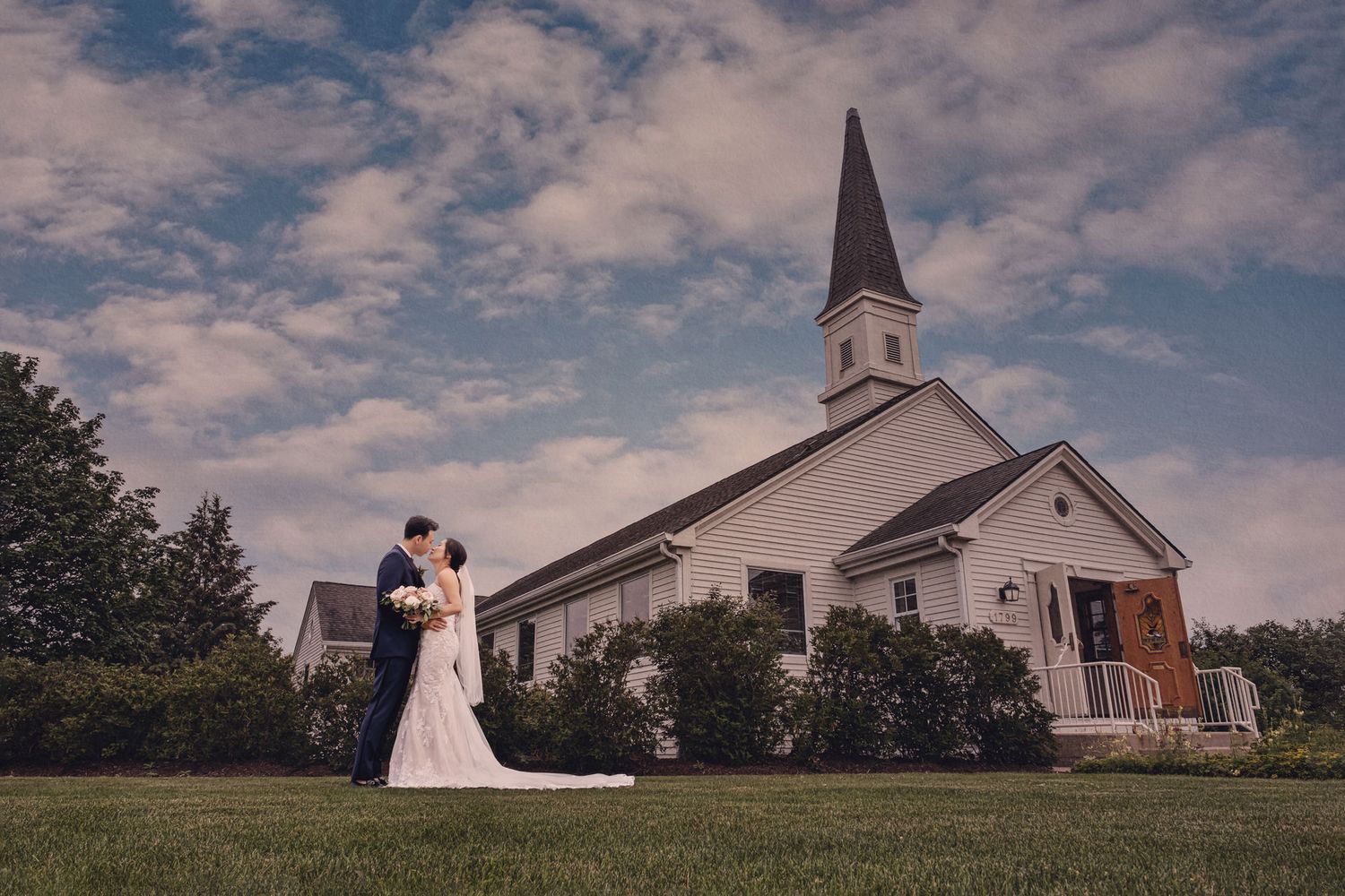 A classic white church with a tall steeple stands against a cloudy sky while a couple shares a romantic moment on the lawn.