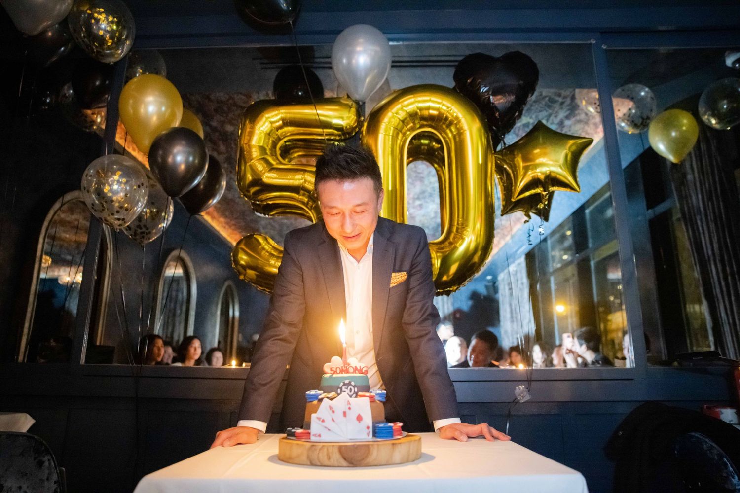 Celebratory scene with gold number 50 balloons, party decorations and a birthday cake with glowing candles on white tablecloth.