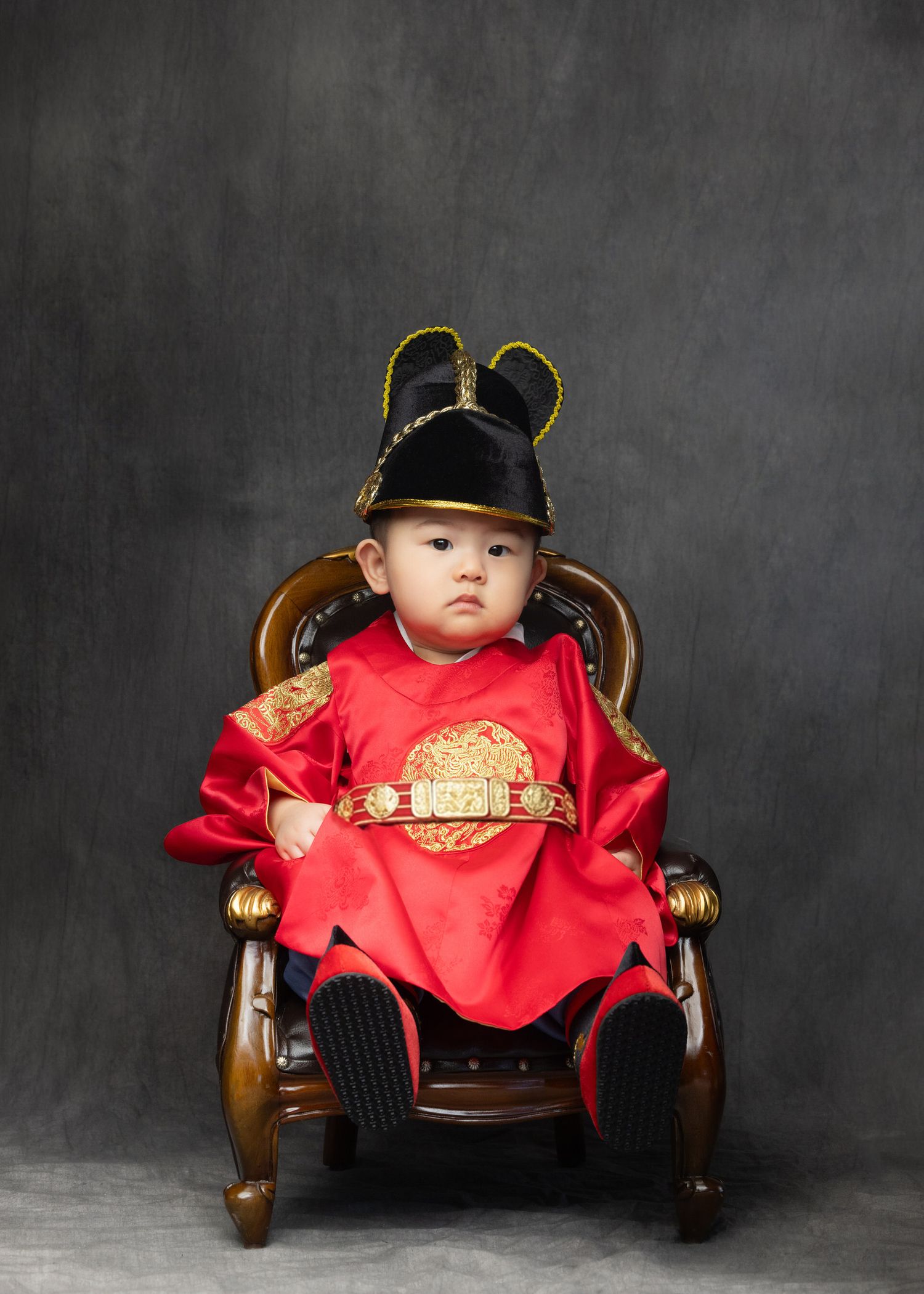 A small child in traditional Chinese New Year attire sits in an ornate wooden chair against a dark backdrop.