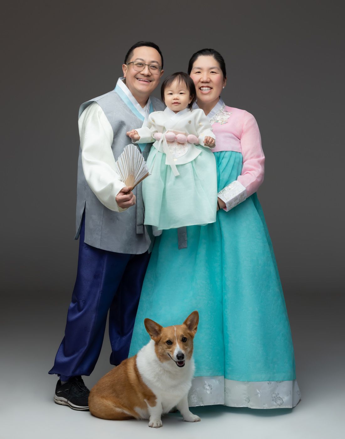 A family wearing traditional Korean hanbok poses together with their Corgi dog in a studio portrait series.