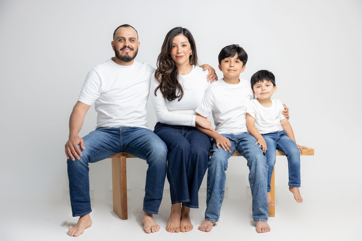 Family of four in white shirts and jeans sitting together on a wooden bench against a white background.