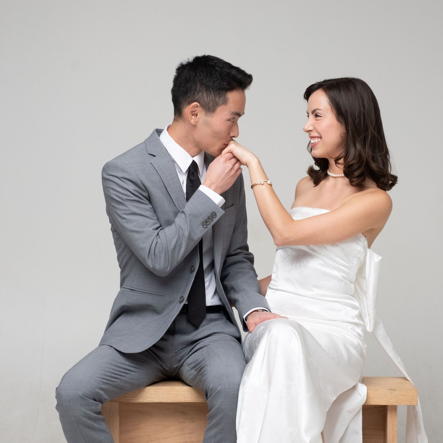 Happy couple in wedding attire laughs together while seated on wooden chairs against gray background.