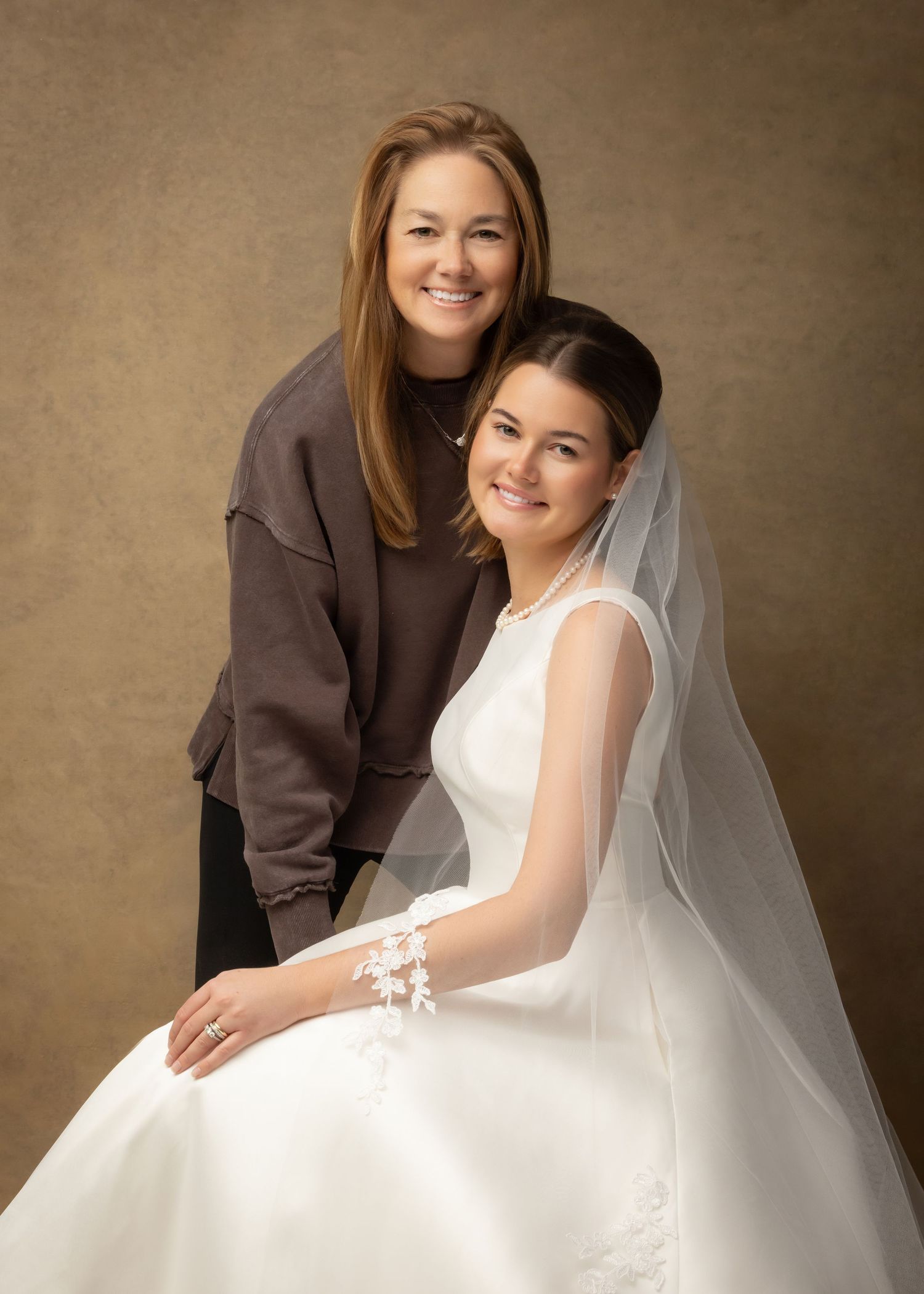 A young girl in a white First Communion dress and veil poses with a smiling woman in a brown sweater.