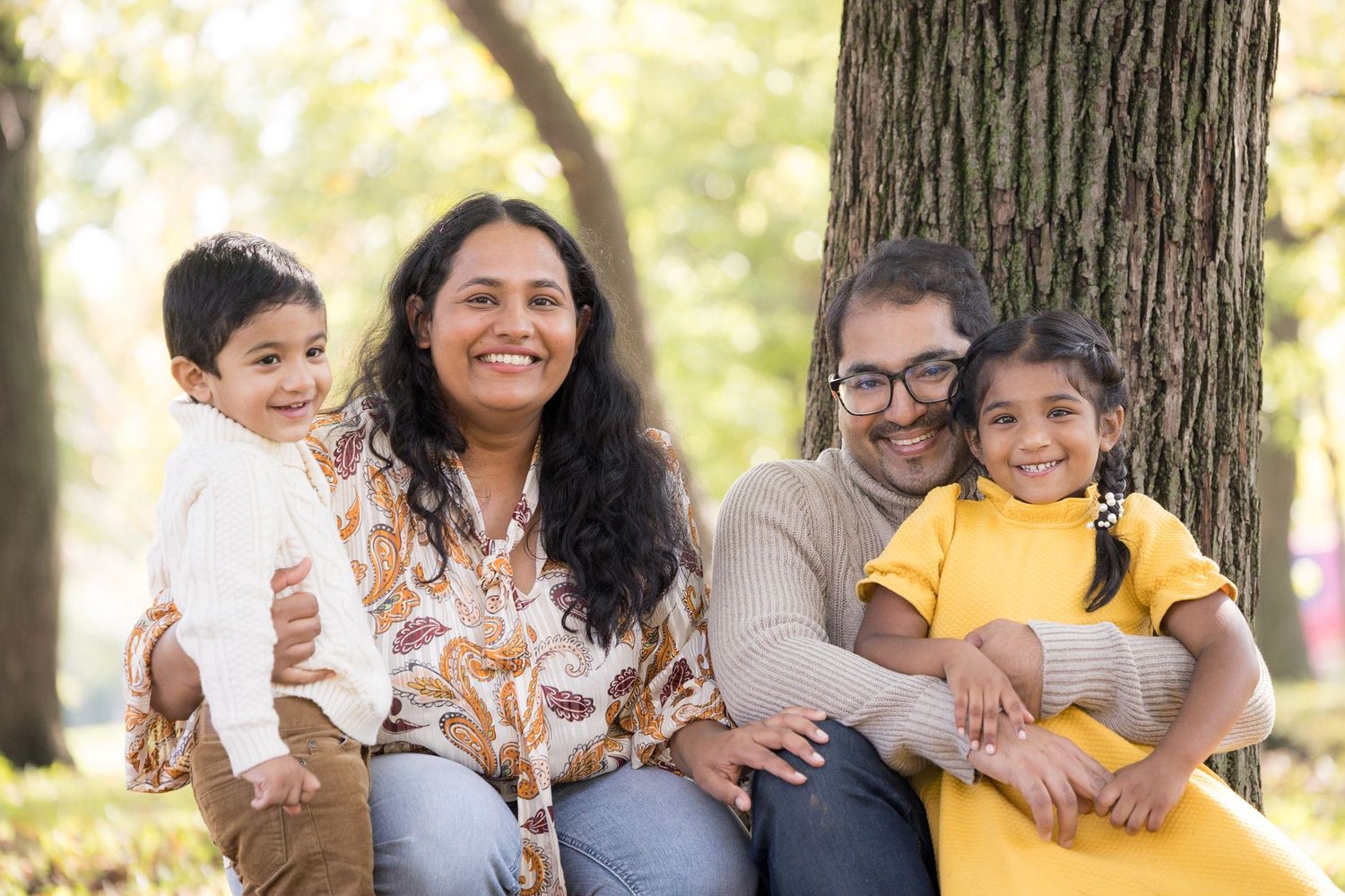A family sits together outdoors against a tree trunk on an autumn day.