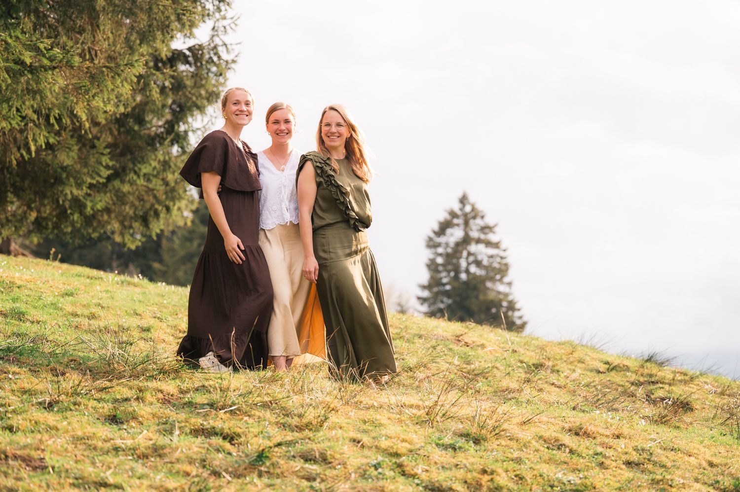 Three women in elegant maxi dresses posing together on a grassy hillside with evergreen trees in background.