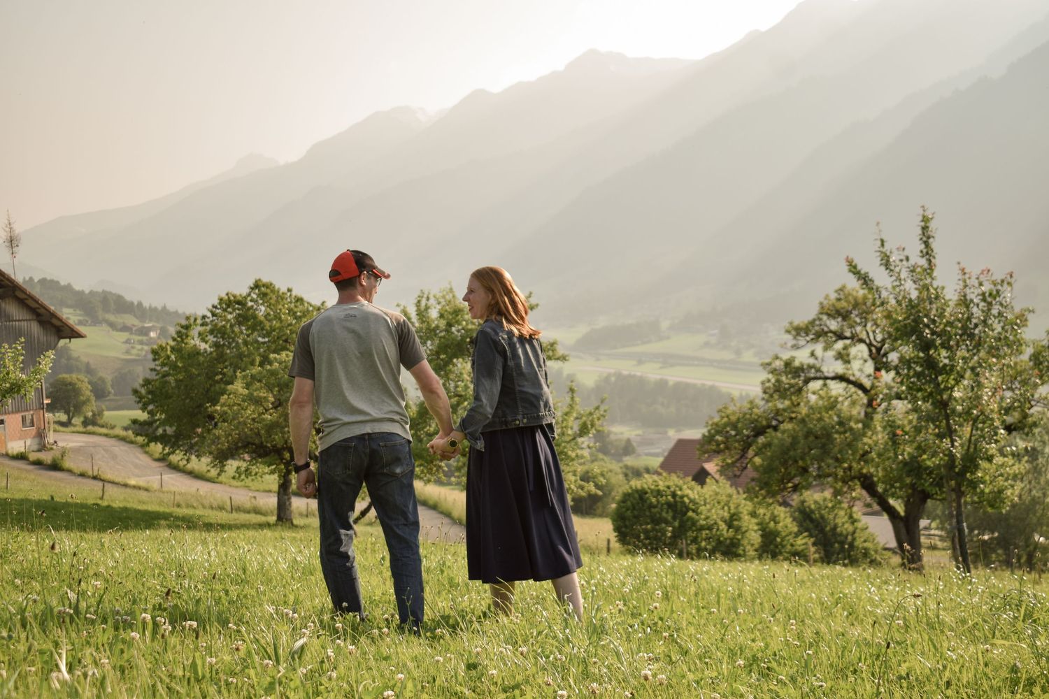 Panoramic view of a couple walking in a green meadow with mountains in the background.