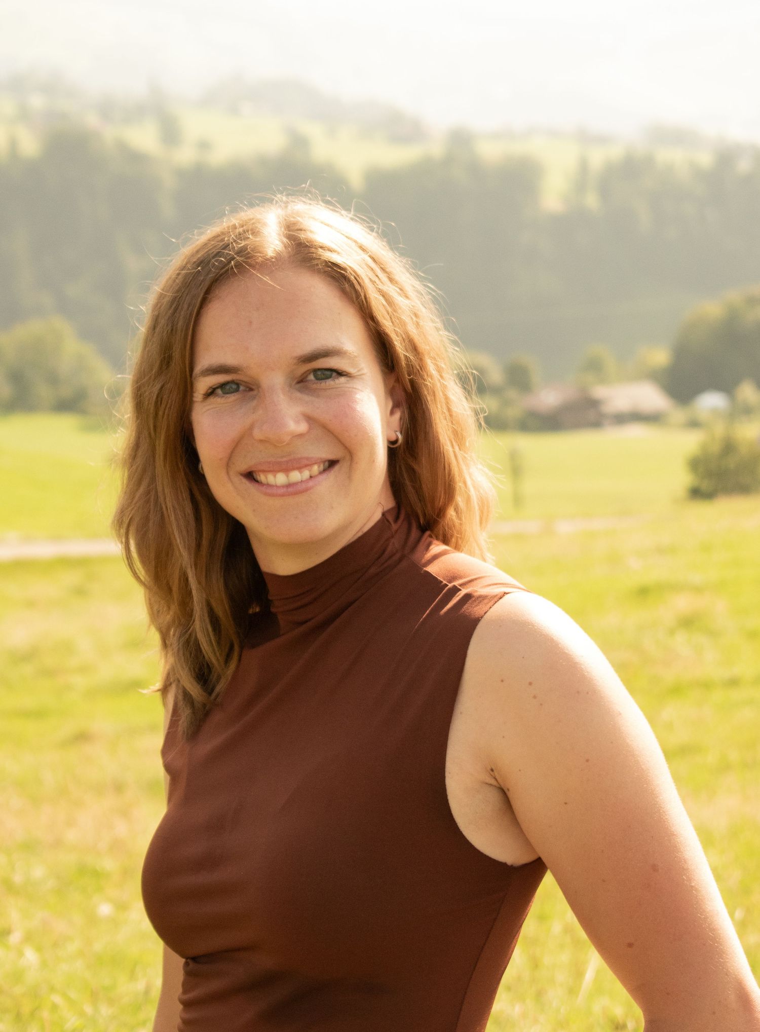 Person in brown sleeveless top smiling in sunny mountain meadow.