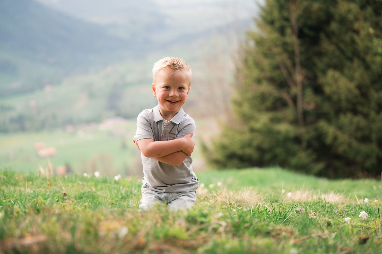 Smiling blonde boy with arms crossed standing on a green hillside with trees and mountains in the background.