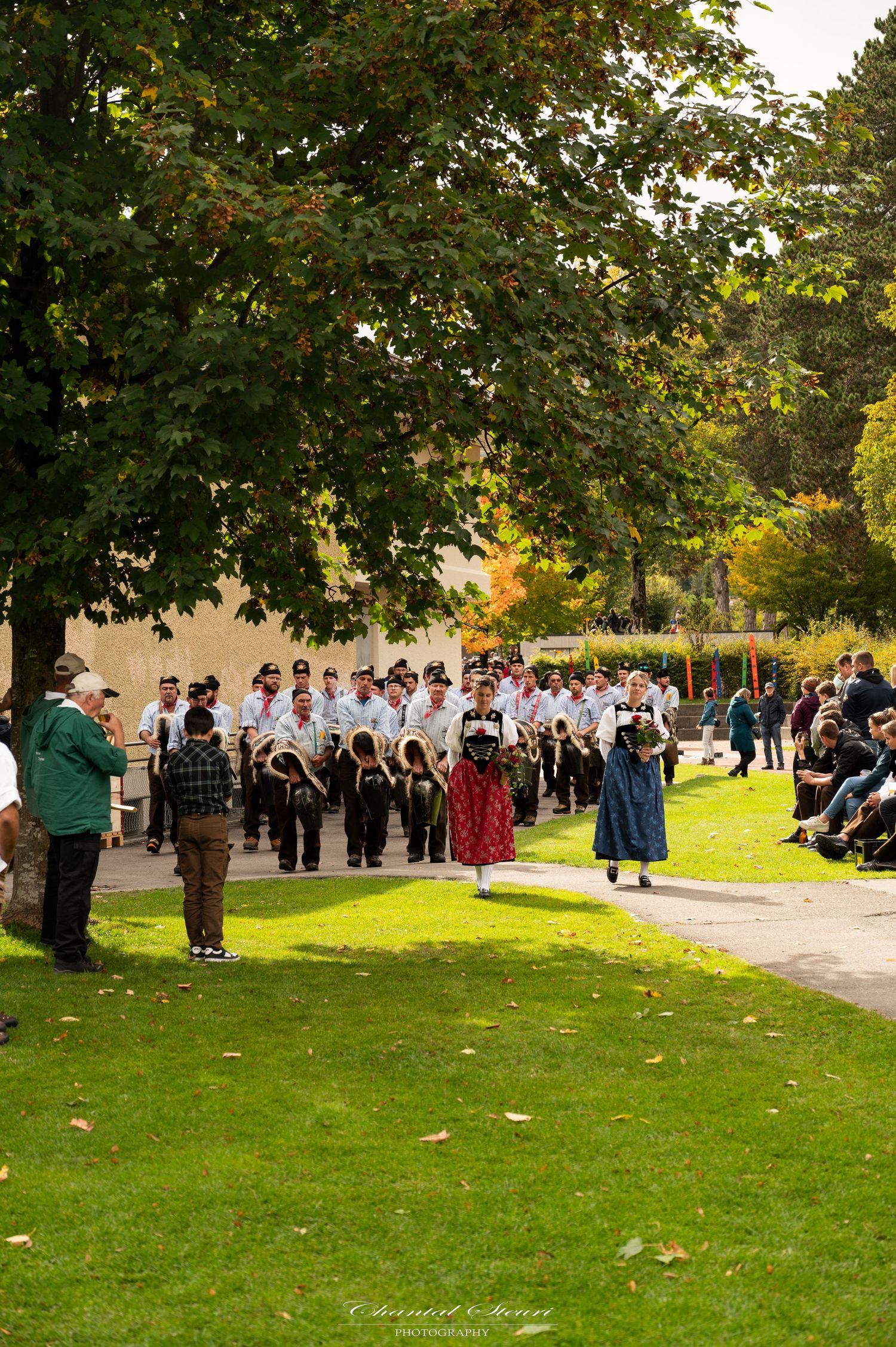 A large outdoor gathering of people in formal attire standing on a grassy lawn during a sunny day.