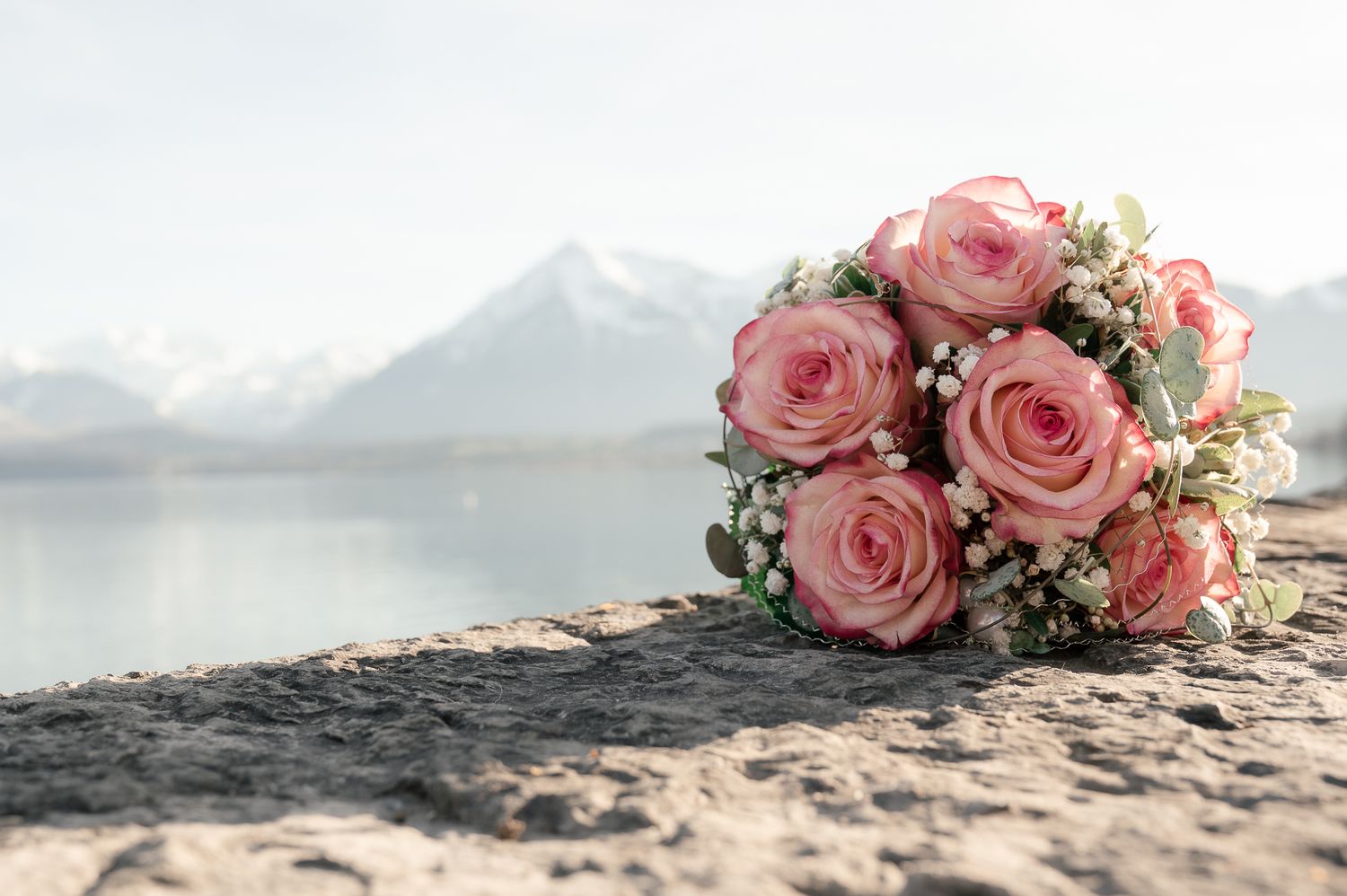 Pink rose wedding bouquet rests on rocky surface with misty mountain lake view in background.