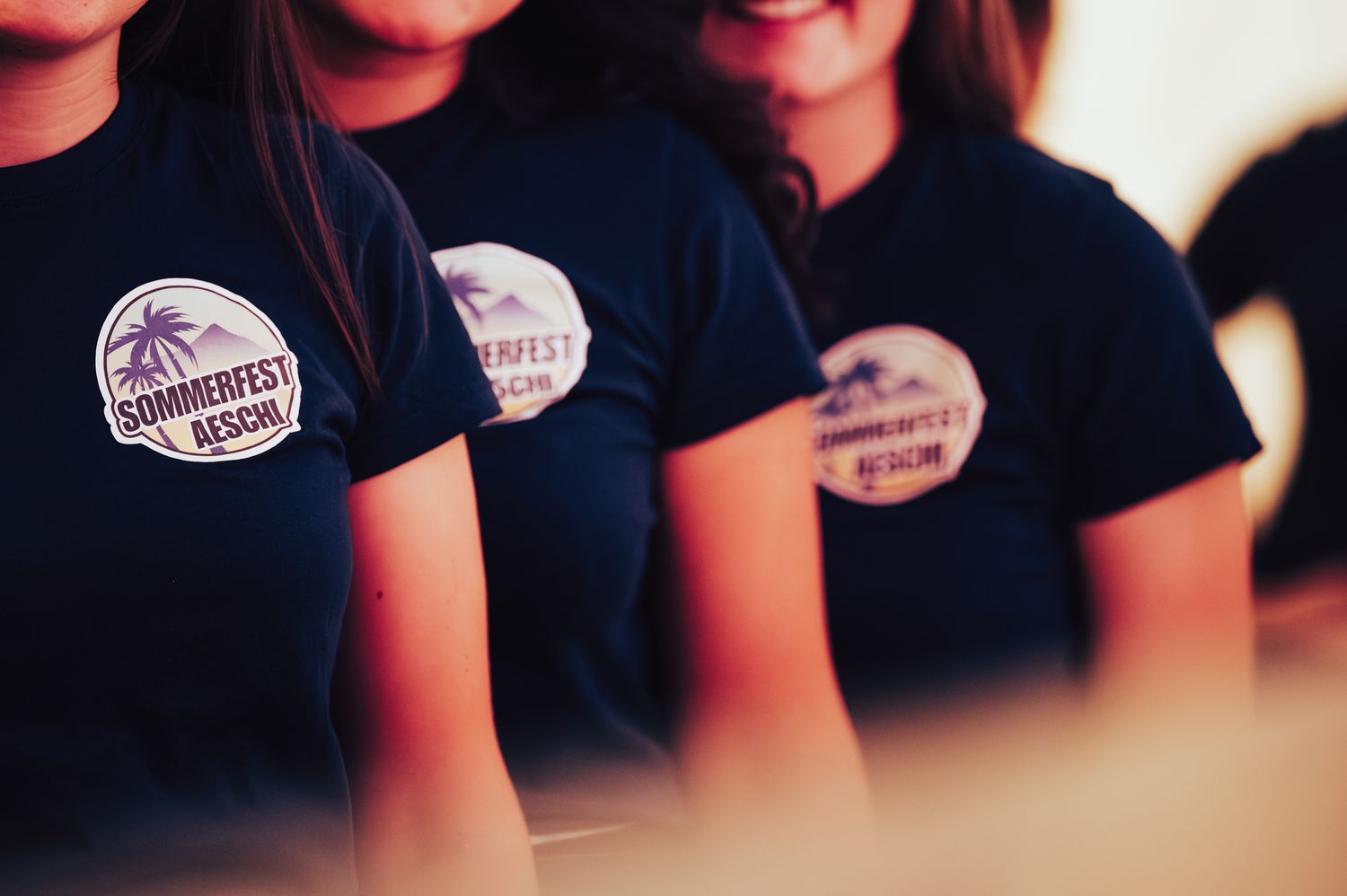 Three women wearing matching navy blue Sommerfest t-shirts with circular badges smile at an outdoor event.