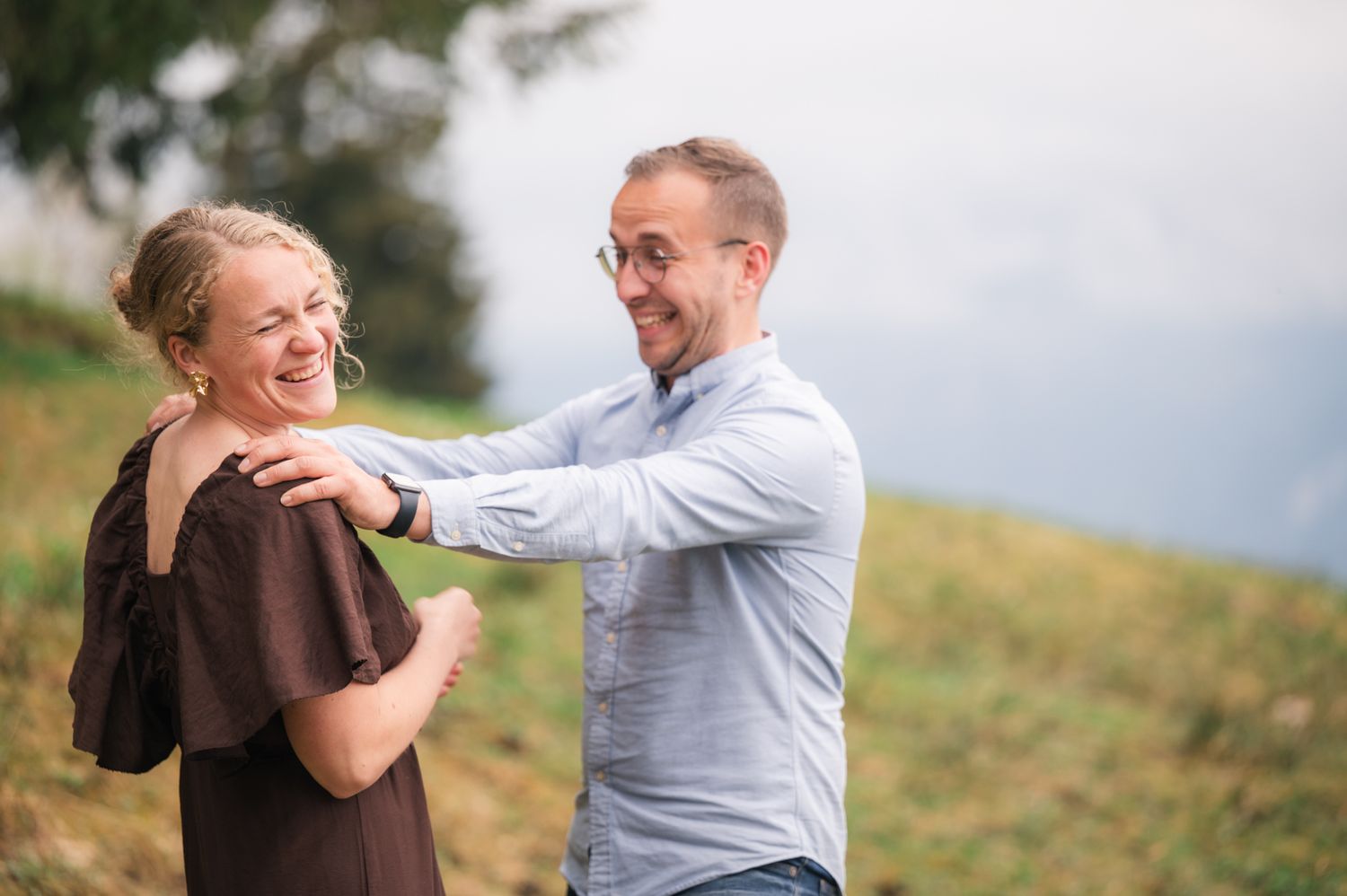 A smiling couple laughing together outdoors on a grassy hillside, man in blue shirt with glasses touching woman's shoulder.
