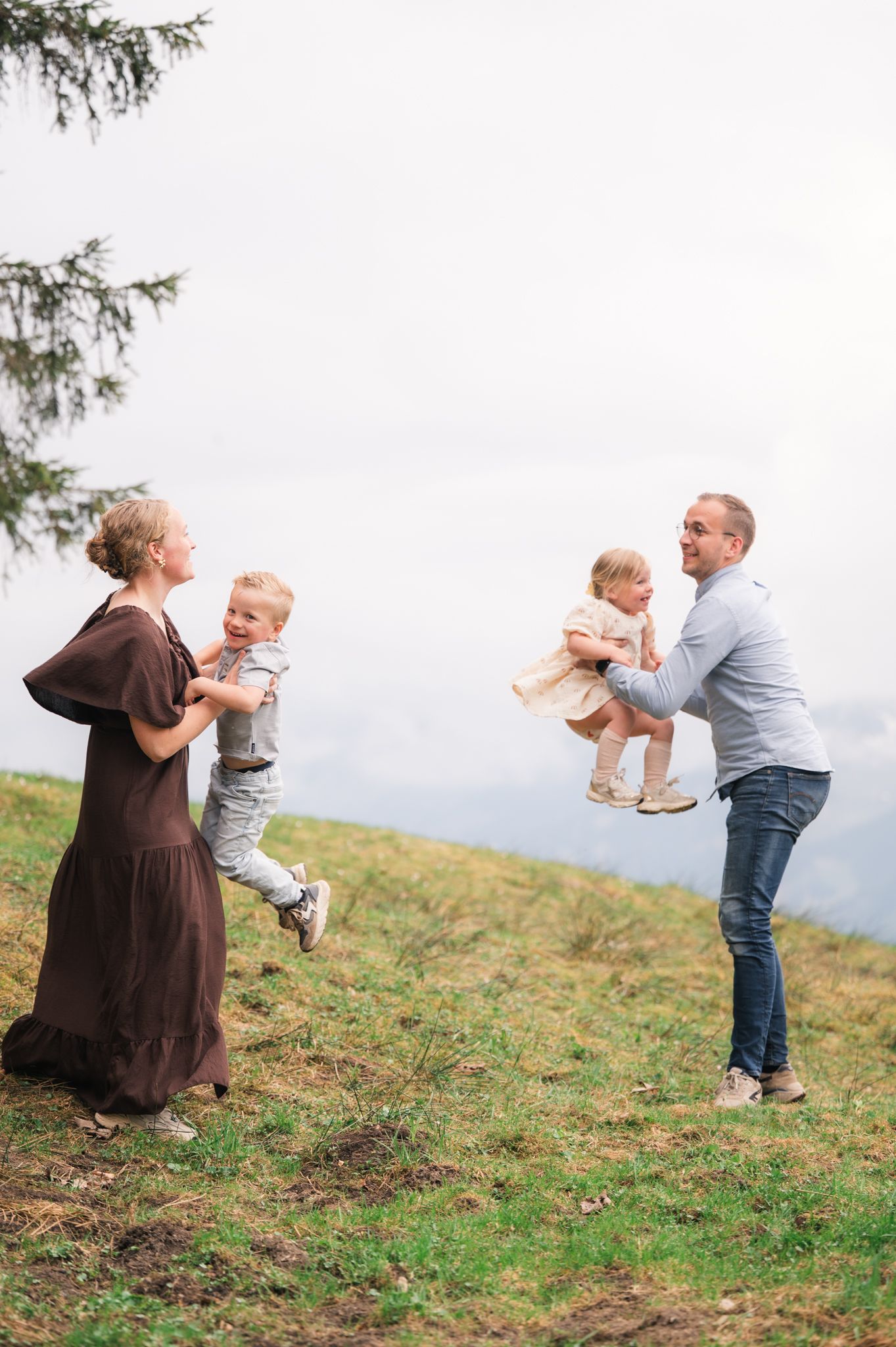 Family of four playing outdoors on a grassy hillside, parents lifting two toddlers joyfully.
