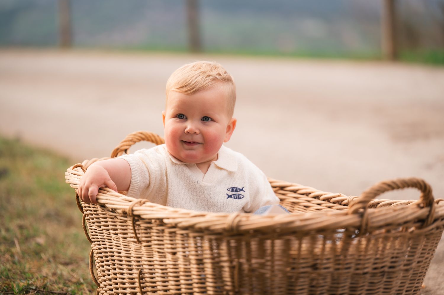 Smiling baby boy sitting in a large wicker basket outdoors on a sunny day.