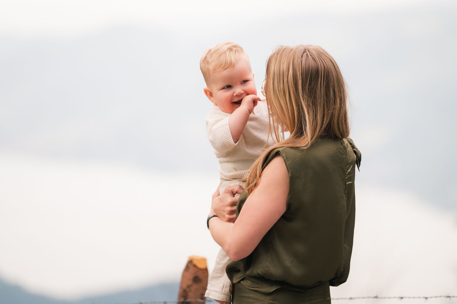 A mother holds her smiling baby outdoors with a scenic mountain lake backdrop.