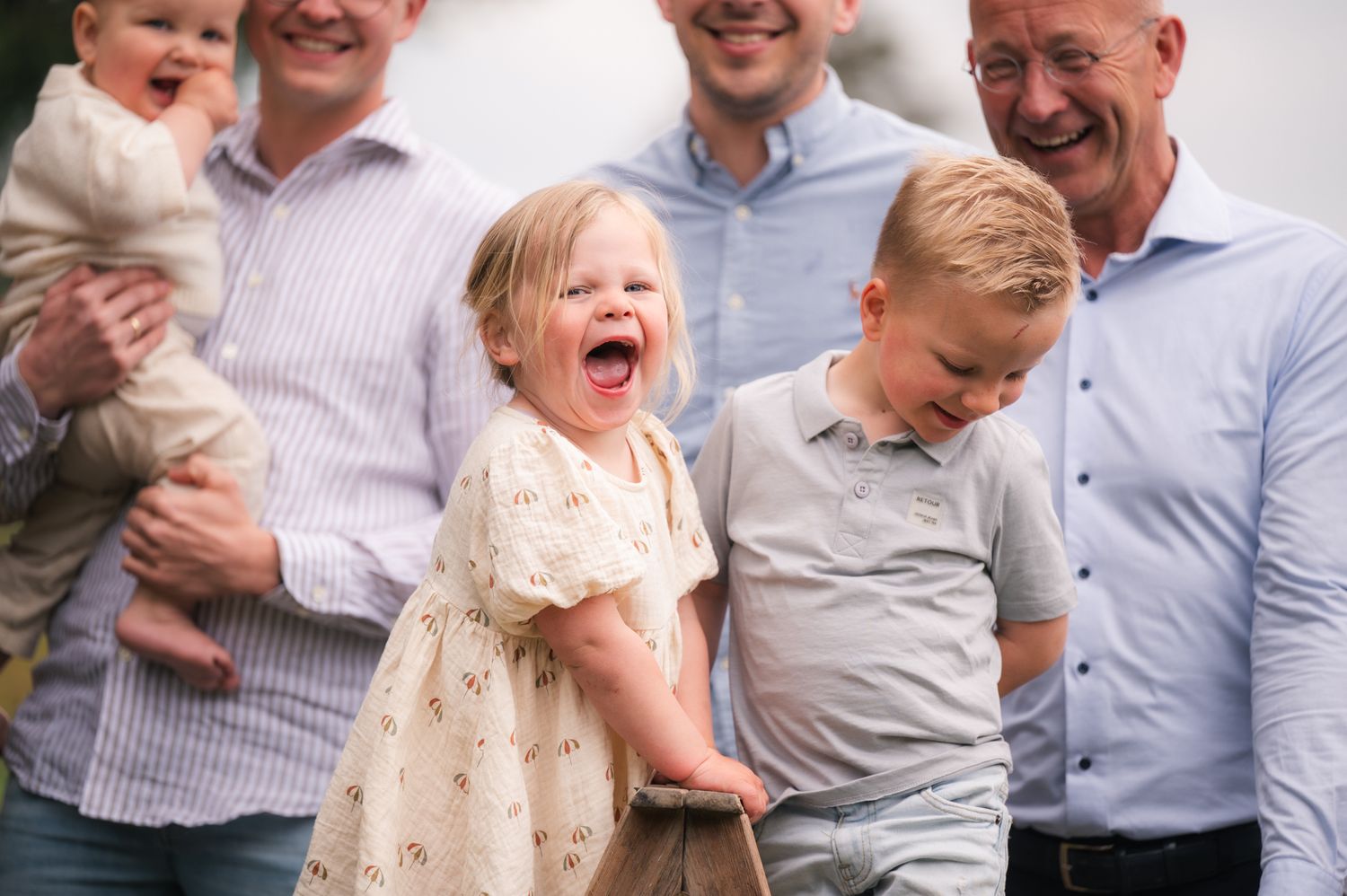 Joyful toddler girl laughing outdoors with family, including two men and a young boy, during a candid family photo session.