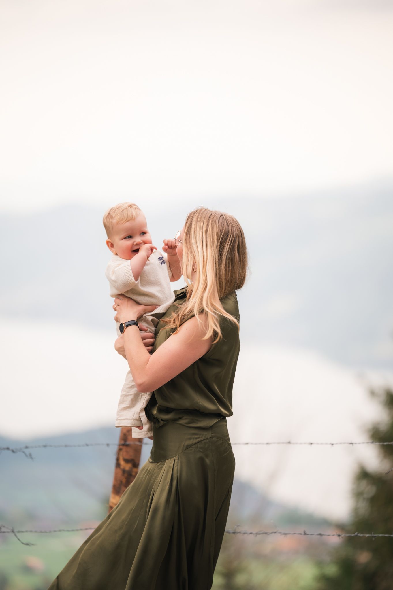 A blonde woman in a green dress holds a baby up while standing outdoors with mountains in the background.