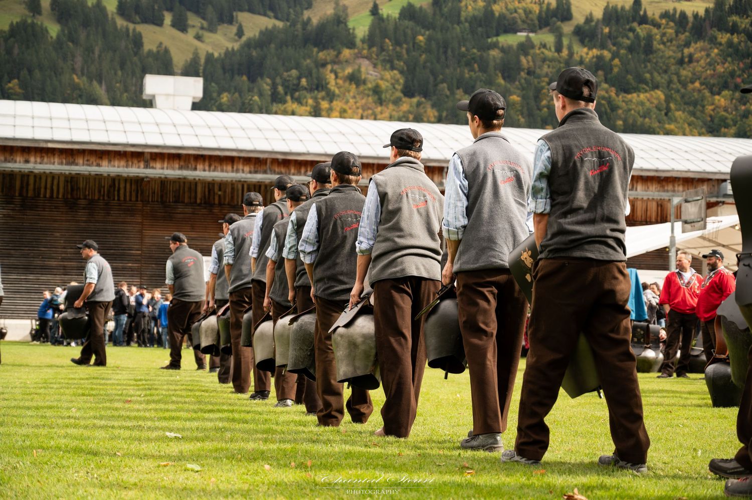 Line of traditionally dressed performers carrying milk cans across a grassy field.