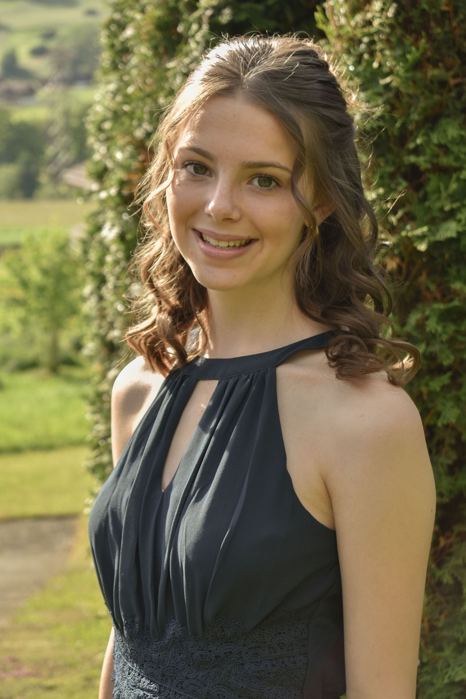 Woman in a black sleeveless dress poses outdoors against green foliage.