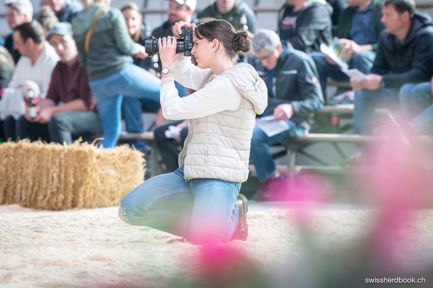 Woman photographer kneeling with camera at outdoor livestock event, hay bales and crowd visible in background.