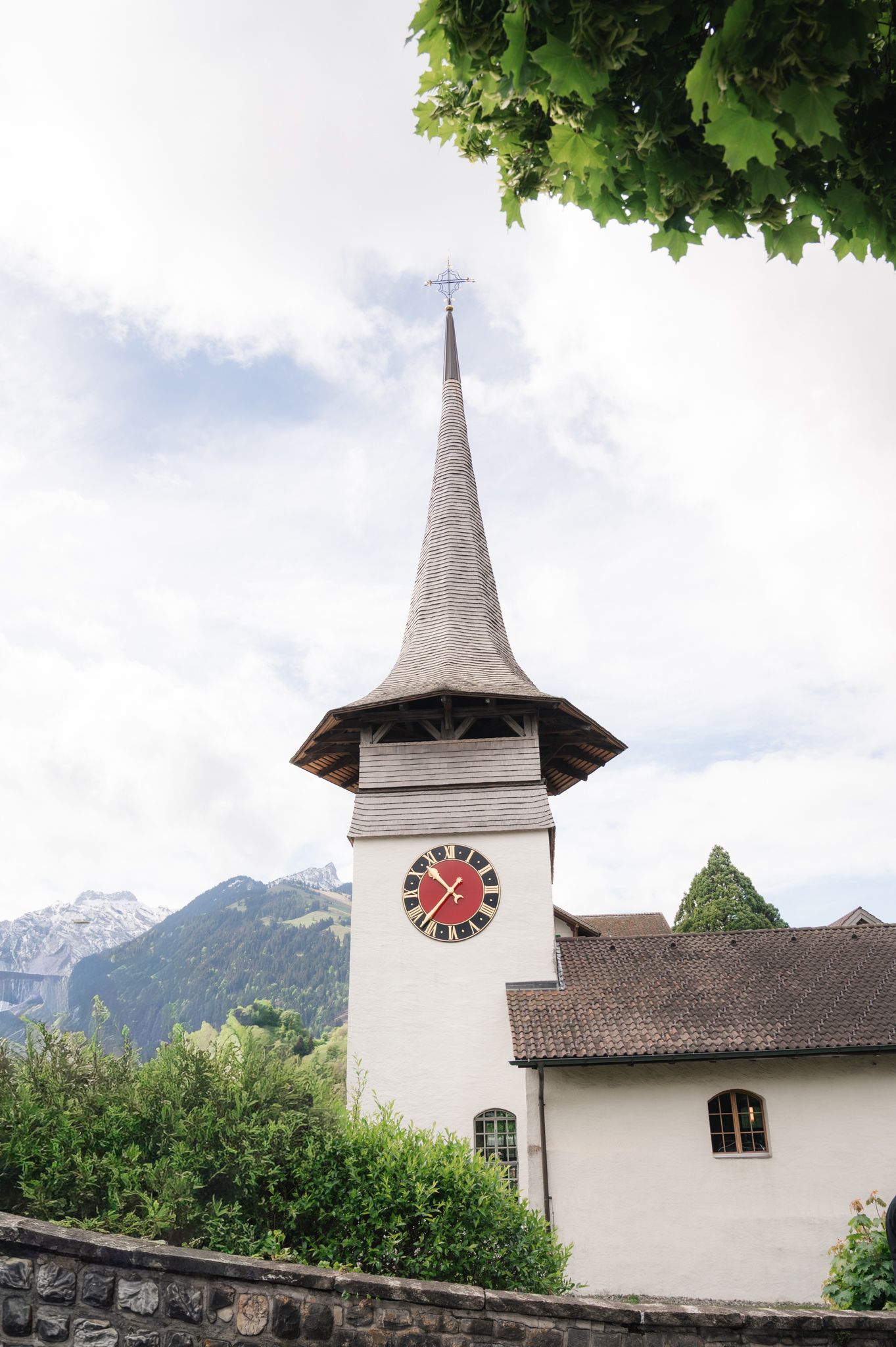White church clock tower with red clock face and pointed spire, set against mountains and cloudy sky.
