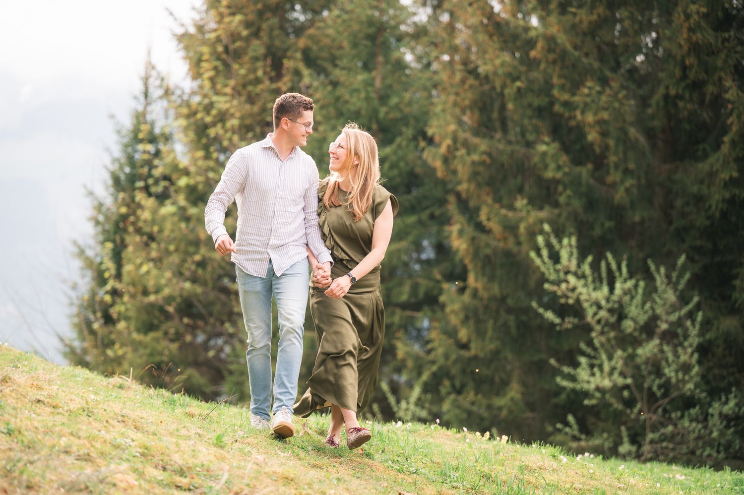 Couple walking hand in hand on a grassy hillside surrounded by tall evergreen trees on a cloudy day.