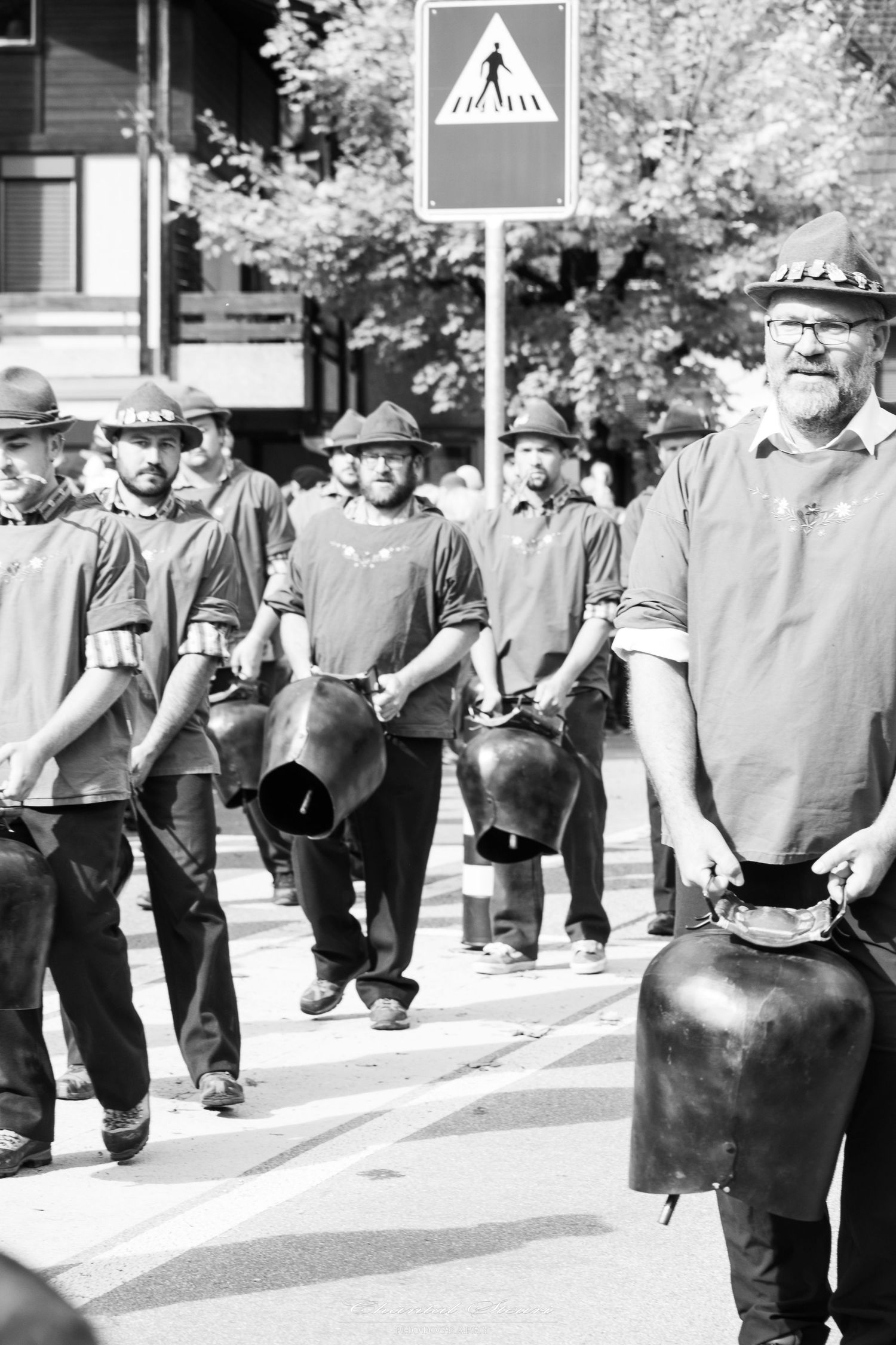 Group walking in line carrying traditional milk cans past a crosswalk sign.