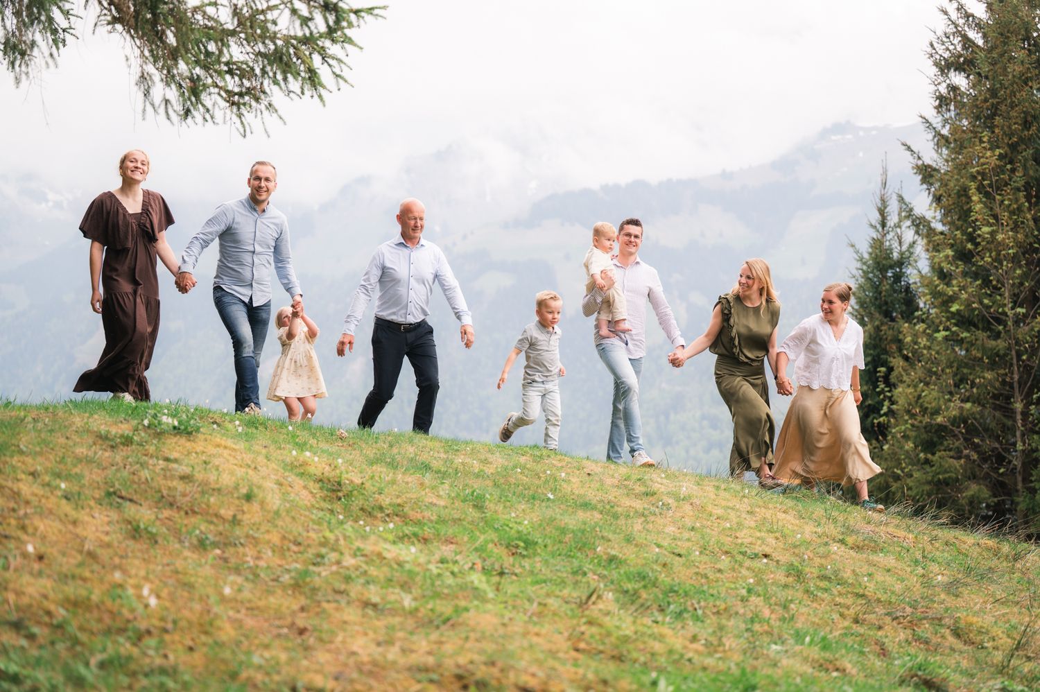 Large family holding hands walking on a green hillside with mountain views and pine trees in background.