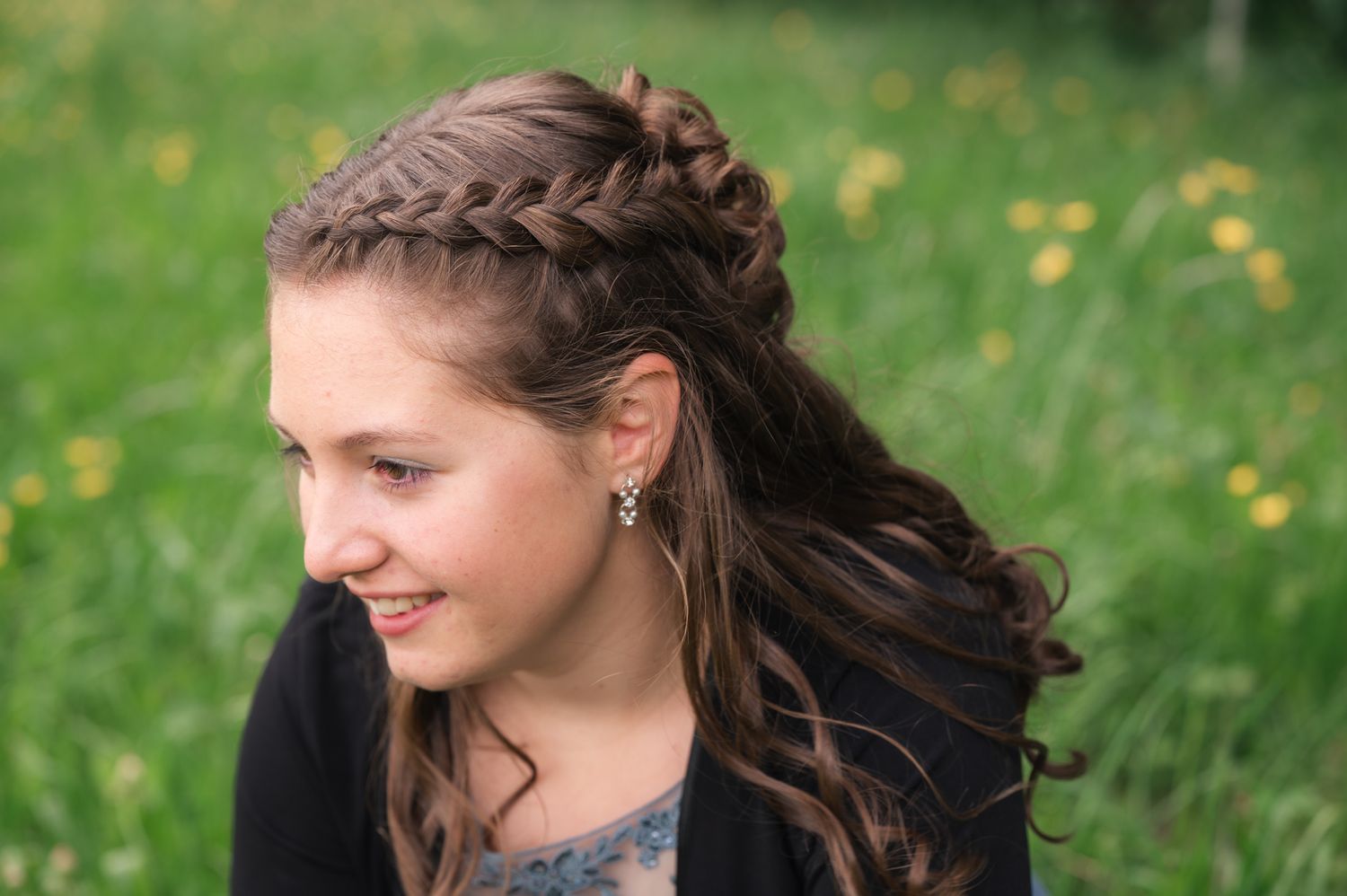Young girl with a braided half-updo hairstyle smiling in a green meadow with wildflowers.