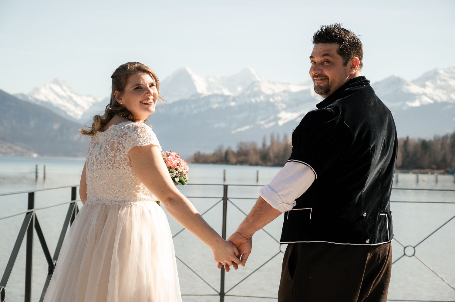 A couple shares a romantic moment on a dock overlooking a snow-capped mountain lake on their wedding day.