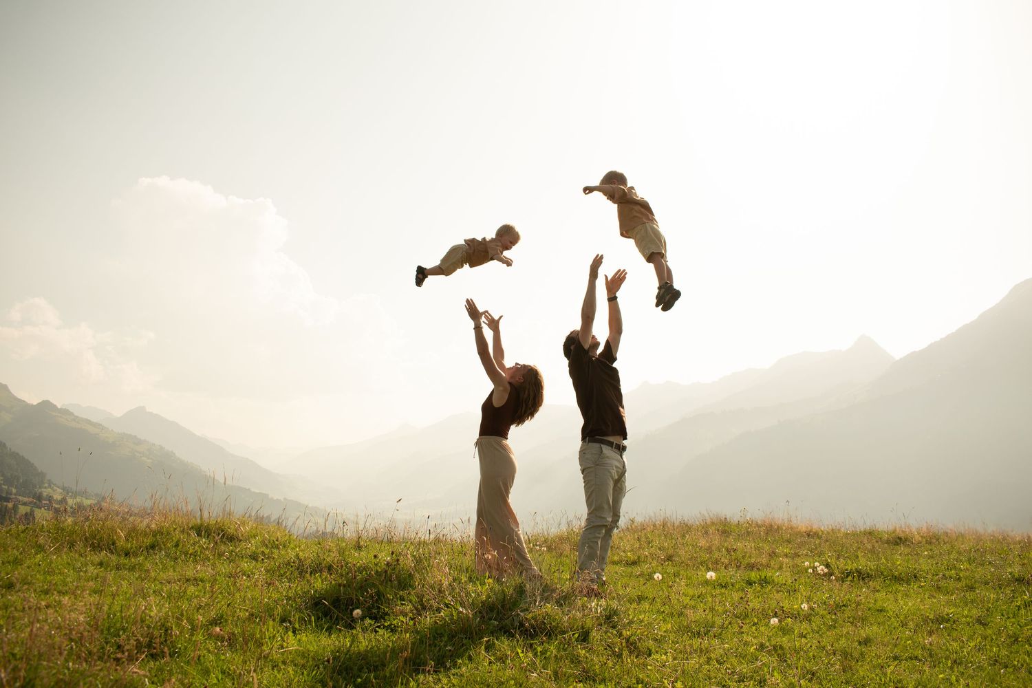 Family joyfully tossing two children in the air on a sunny mountain meadow with scenic Alpine peaks in background.