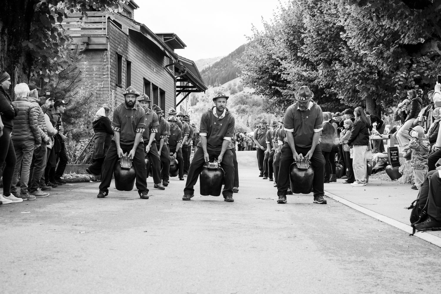 A vintage black and white photograph showing a group of people walking down a village street carrying luggage.