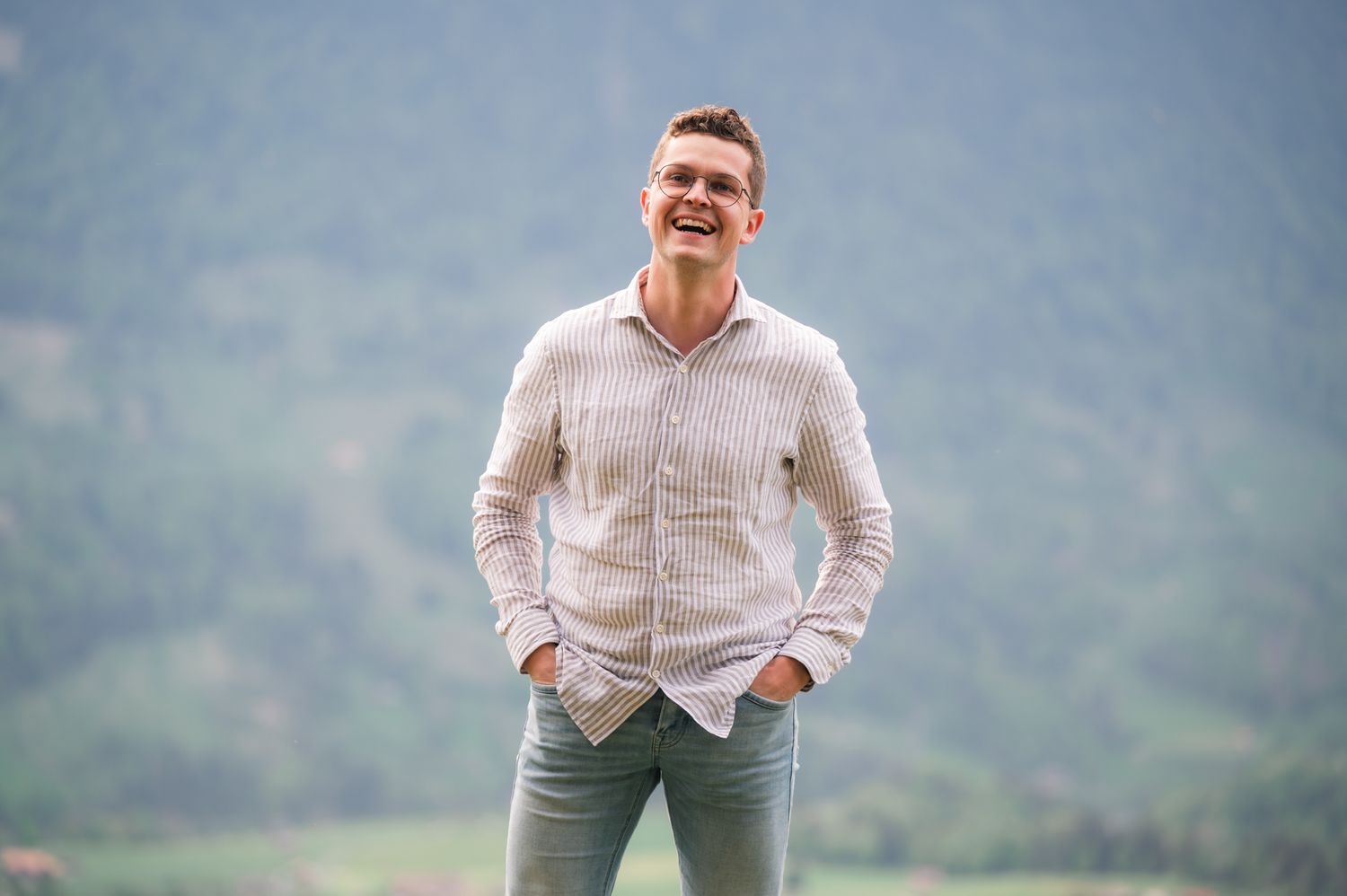 Young man smiling outdoors with hands in pockets, wearing light shirt and green pants, green hills in background.