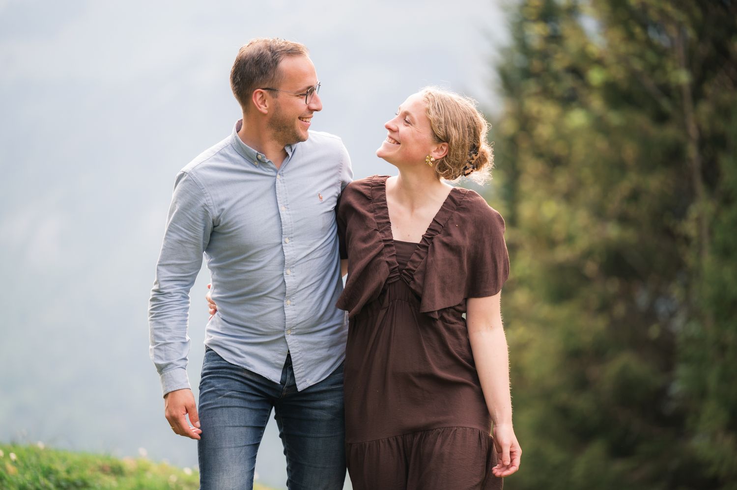 A smiling couple walks together outdoors, with lush green trees in the background on a cloudy day.
