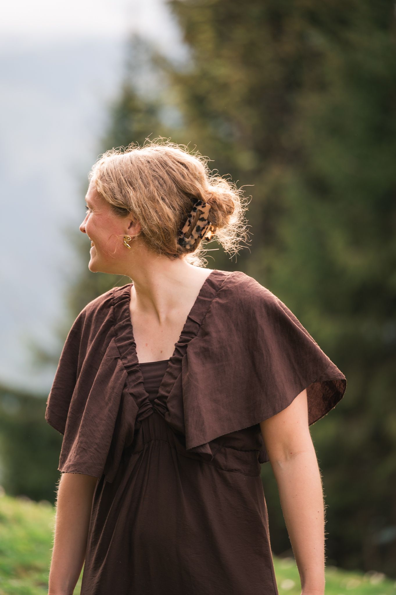 Young woman in brown dress with braided updo smiling outdoors with green trees in background.