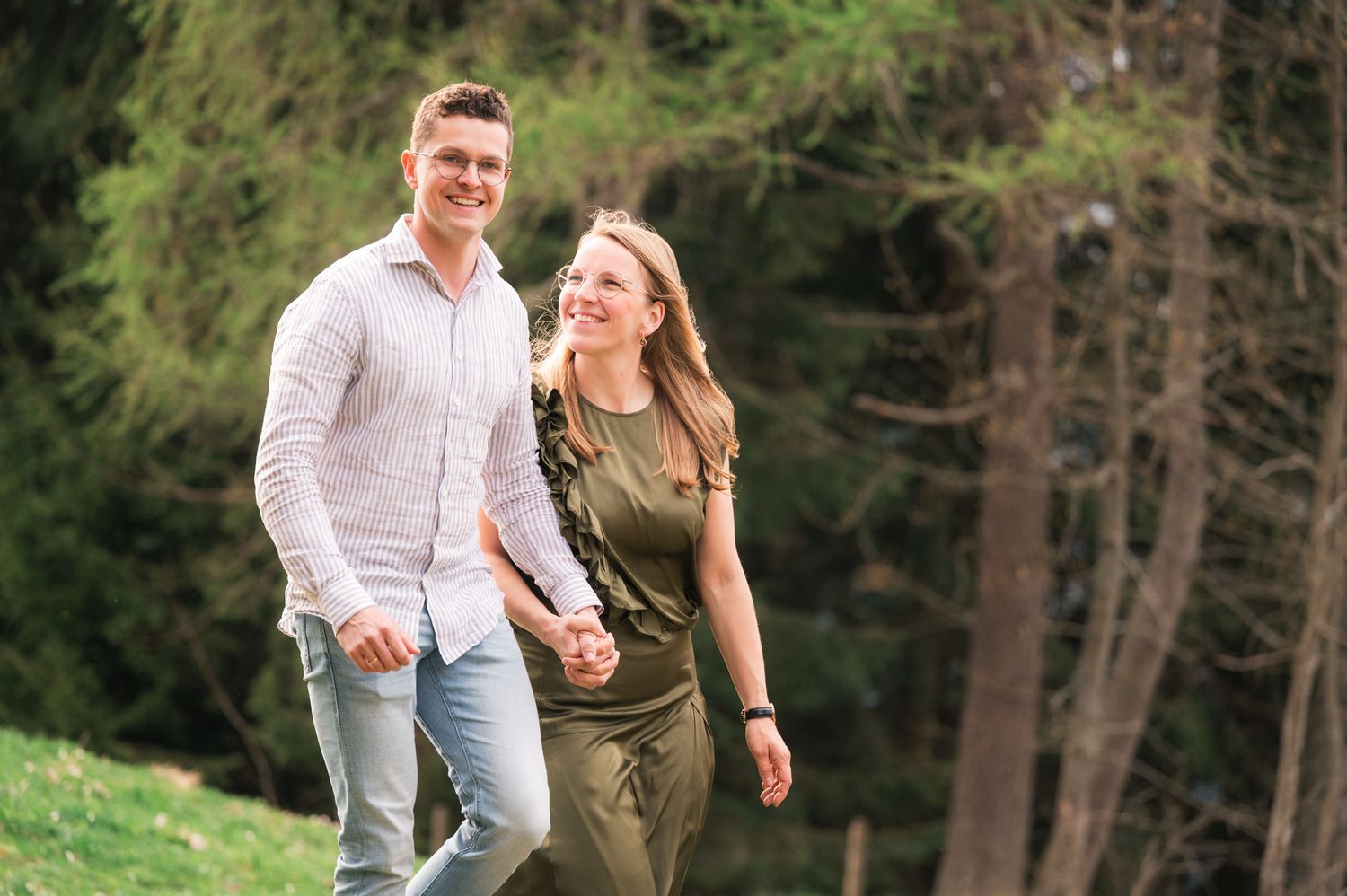 Happy couple holding hands while walking outdoors in a lush green forest setting on a sunny day.