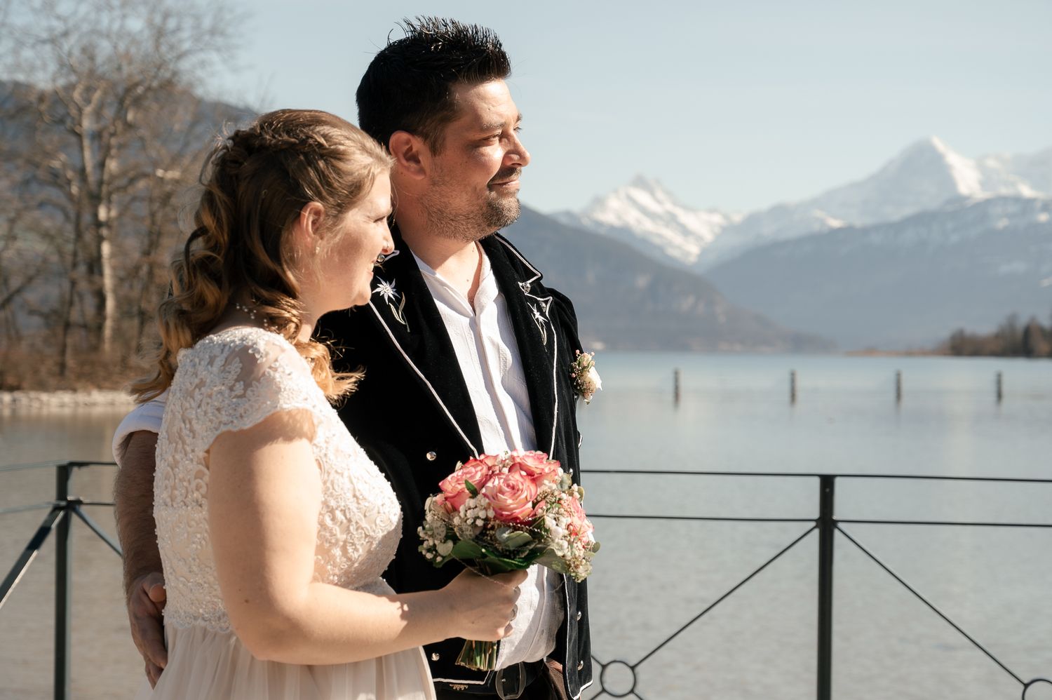 A couple in wedding attire embrace while standing near a scenic mountain lake with snow-capped peaks in the background.