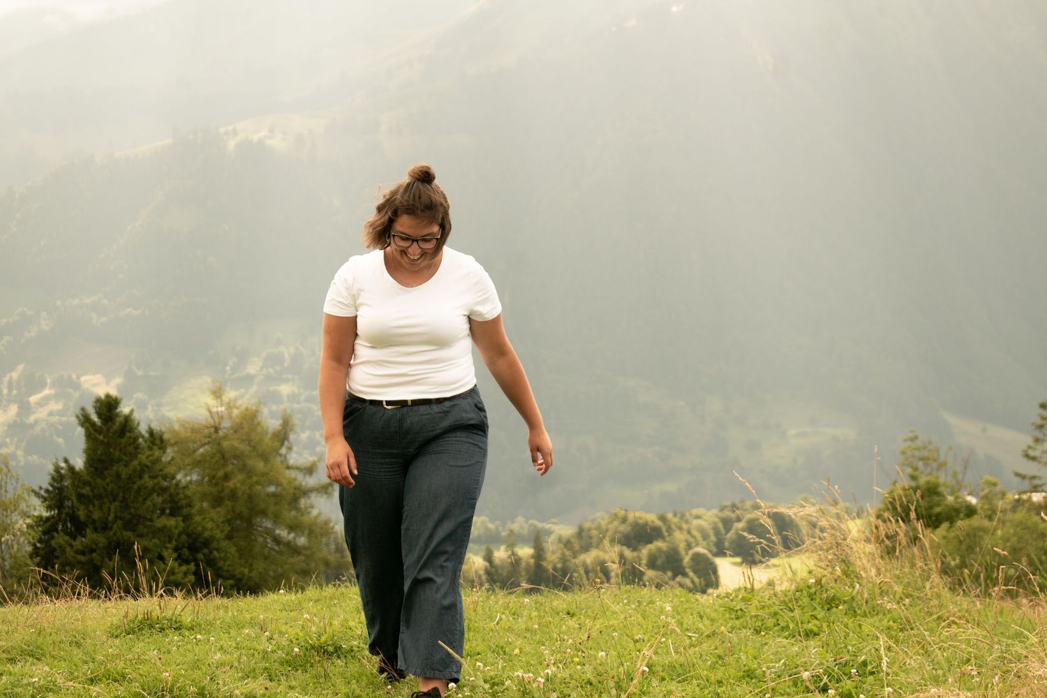 Person walking on grassy mountain path wearing white top and dark pants with scenic valley below.