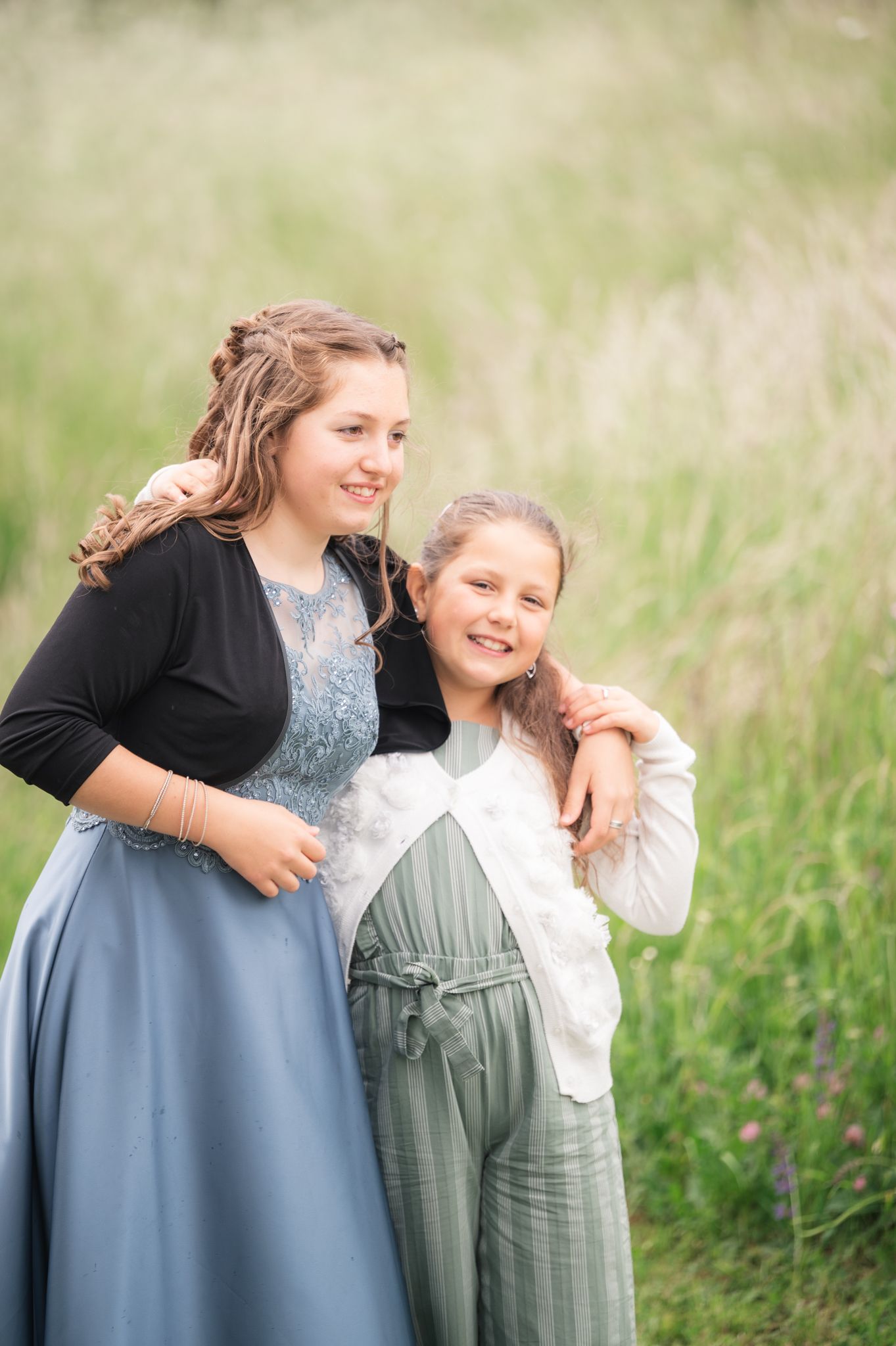 Two smiling girls in elegant dresses pose together in a lush green field for a portrait photo.