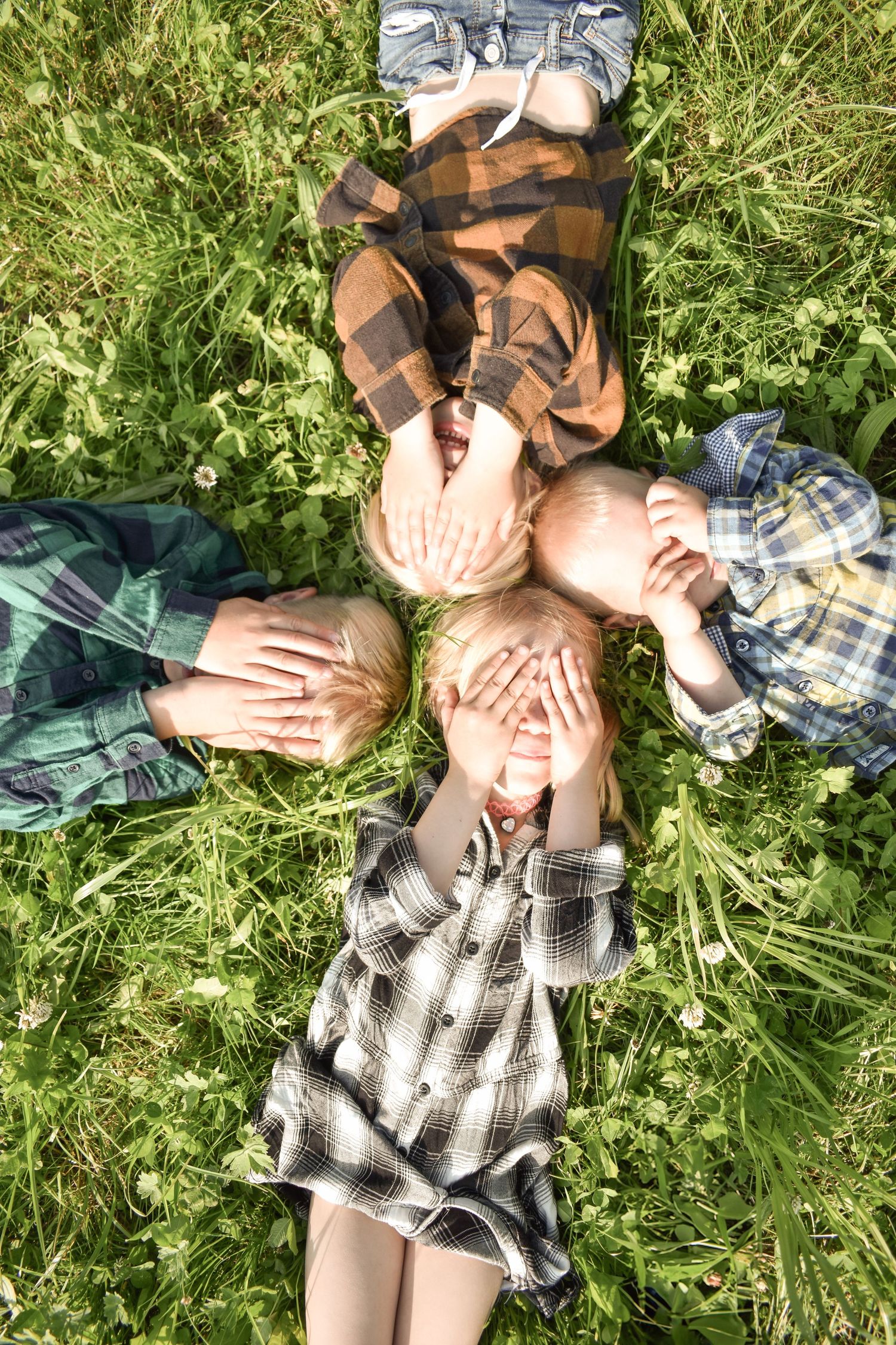 Four people lying in grass wearing plaid shirts playfully covering their faces with hands.