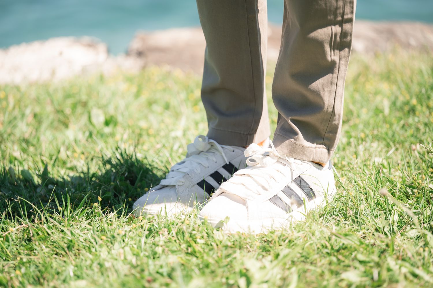 Person standing on green grass wearing white Adidas Superstar sneakers with black stripes and khaki pants.